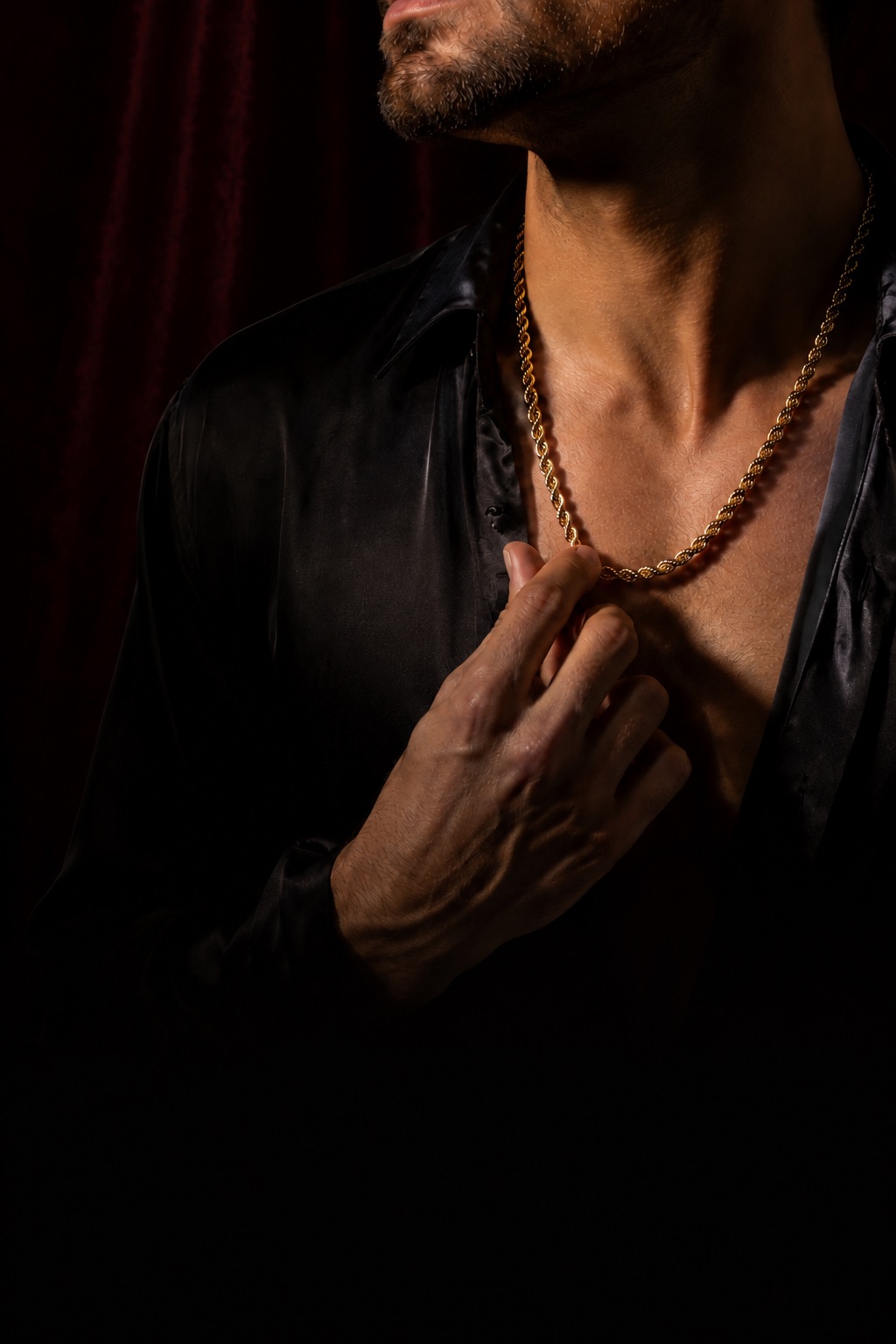 A man in an open black shirt wearing a heavy gold rope chain, lit against a deep red curtain.